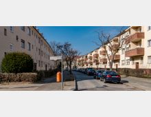 Wohnstraße mit mehrstöckigen Wohnhäusern aus hellem Putz und roten Klinkerfassaden, die auf beiden Seiten der Straße stehen. Geparkte Autos säumen die gepflasterte Straße, und kahle Bäume stehen entlang des Gehwegs; ein Straßenschild mit der Aufschrift „Deutsche Straße“ ist sichtbar.