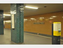 U-Bahn-Station mit gelb-orangefarbenen, gefliesten Wänden und türkisfarbenen Säulen. Ein Schild mit der Aufschrift "Güntzelstraße" ist an der Wand angebracht; im Vordergrund ein gelber Automaten für Fahrkarten.