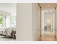 Helles, modernes Schlafzimmer mit einem Bett, das in Richtung eines Balkons mit großen Glastüren ausgerichtet ist, neben einem schmalen Flur mit Einbauschränken und Holzfußboden.