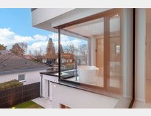 Moderne Wohnhausfassade mit bodentiefen Glasfronten und Holzrahmen, die einen hellen Innenraum mit freistehender Badewanne zeigen. Im Hintergrund sind ein Wohngebiet mit Ziegeldächern, Gartenzaun und Bäumen unter blauem Himmel zu sehen.
