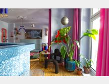 Bright, colorful living room with large windows framed by pink curtains, a wooden floor, and several potted plants including a tall leafy plant in a blue pot. The space features playful modern decor such as sculptural black seating, a small round side table, hanging globe lights, and wall art in pastel tones.
