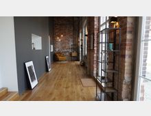 Industrie-inspirierter Innenraum mit Backsteinwänden, großen Fenstern und einem Holzboden. An der linken Wand lehnen gerahmte Bilder, während rechts ein schmales Regal steht; eine gelbe Sesselgruppe ist im Hintergrund sichtbar.