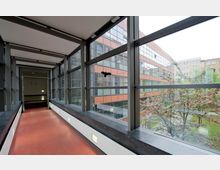 Ein moderner überdachter Verbindungsgang mit großen Fensterflächen und Metallrahmen, der auf einen Innenhof mit blühenden Bäumen und umliegenden Bürogebäuden blickt. Der Boden des Gangs ist rot, und die Fenstersimse sind aus dunklem Stein.