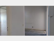 Leerer Raum mit weißen Wänden und grauem Boden, teilweise sichtbare Glastür auf der linken Seite mit geriffeltem Glas, rechts ein Türrahmen zu einem weiteren Raum.