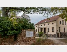 Ein zweigeschossiges, beigefarbenes Gebäude mit rotem Ziegeldach und mehreren Fenstern, flankiert von einem Backsteinzaun mit Informationen auf einer Tafel. Davor befinden sich Bäume und Büsche, die den Eingang teilweise umrahmen.