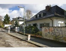 Wohnstraße mit freistehenden Einfamilienhäusern, umgeben von Zäunen und Gärten. Im Vordergrund ein weißes Haus mit dunklem Dach und vergitterten Fenstern, daneben eine Mauer mit Graffiti; Gehweg und Laternen säumen die Straße.