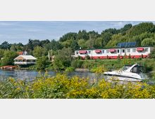 Flusslandschaft mit einem kleinen, modernen Gebäude mit roten Markisen im Hintergrund und einer Holzterrasse mit Überdachung am Ufer. Im Vordergrund liegt ein Motorboot auf dem Wasser, umgeben von grüner Vegetation und gelben Blumen.