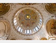 Innenansicht einer reich verzierten Kuppel einer Kirche. Die Decke ist mit Gemälden, goldenen Ornamenten und Reliefs gestaltet, während große Fenster Licht einlassen und die Gestaltung betonen.