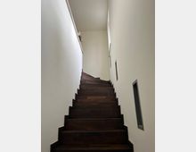 Holztreppe in einem modernen Innenraum mit weißen Wänden, schmalem Aufgang und rechteckigen Wandnischen auf der rechten Seite. Oben befindet sich ein Fenster, das Tageslicht hereinlässt.