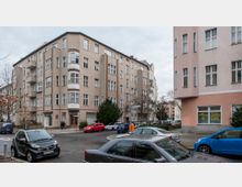 Straßenszene mit mehrstöckigen Wohnhäusern im typischen Stil der frühen 20. Jahrhunderts, hellbraune und rosa Fassaden mit zahlreichen Fenstern. Im Vordergrund parkende Autos und ein kleiner Platz mit Bäumen und Gehwegen.