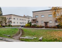 Wohnsiedlung mit Flachdachgebäuden im Stil der Moderne, umgeben von kleinen Gärten, Gehwegen und einer Grünfläche im Vordergrund. Die Fassaden sind in beige gehalten, mit roten Backsteindetails an Sockeln und Eingängen, und es sind wenige gepflasterte Wege sowie Herbstlaub sichtbar.