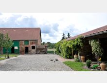 Hof mit Kopfsteinpflaster, umgeben von zwei Backsteingebäuden mit roten Ziegeldächern; das linke Gebäude hat grüne Holztore, während das rechte von Pflanzen und Töpfen dekoriert ist. Im Hintergrund sind Bäume und eine grüne Wiese sichtbar.