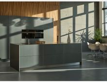 Moderne Küche mit einer freistehenden Kücheneinheit aus mattem Metall, einem integrierten Wasserhahn und einem Holzbrett. Im Hintergrund befinden sich eine Wand mit Holz- und Betonakzenten, eine Reihe von Einbaugeräten, große Fenster mit einfallendem Tageslicht sowie ein Esstisch mit weißen Stühlen und Dekopflanzen.