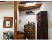 Innenraum mit rustikalen Holzbalken und schrägem Dach mit Oberlicht. Im Raum befinden sich eine Kaffeemaschine, ein Verzierungsspiegel, ein kunstvoll bemalter Schrank und ein großer Holzkleiderschrank mit geschnitzten Details, ergänzend durch kleine Dekorationsobjekte.