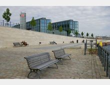 Moderner Flussuferbereich mit Sitzbänken auf einem gepflasterten Gehweg, im Hintergrund ein großes, verglastes Gebäude des Hauptbahnhofs mit einem DB-Logo. Entlang der Uferpromenade stehen vereinzelt Bäume, und Menschen sitzen oder spazieren.