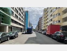 Straßenansicht in einer städtischen Umgebung mit mehrstöckigen Wohn- und Geschäftsgebäuden auf beiden Seiten, teilweise Baustellen mit Baugerüsten und blauer Schutzverkleidung. Geparkte Autos und ein blauer Lkw sowie ein roter Transporter säumen die Straße unter einem bewölkten Himmel.