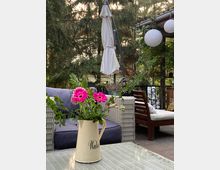 Gartenbereich mit Sitzmöbeln aus Rattan, einem Tisch mit einer cremefarbenen Vase mit pinken Blumen und einem geschlossenen Sonnenschirm im Hintergrund. Rundherum sind Bäume und eine überdachte Pergola mit weißen Papierlampen zu sehen.