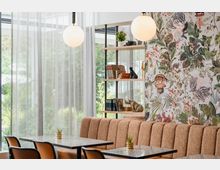 Ein modern eingerichteter Innenraum mit einer gepolsterten Sitzbank in Beige und Marmortischen, umgeben von Stühlen. Im Hintergrund befinden sich durchsichtige Vorhänge vor großen Fenstern, ein Regal mit Büchern und Dekorationsobjekten sowie eine Wand mit farbenfroher Tapete mit Pflanzen- und Tiermustern.