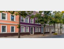 Straßenszene mit mehreren aneinandergereihten Wohnhäusern im historischen Stil, deren Fassaden in Pastellfarben wie Orange und Lila gestrichen sind. Vor den Häusern stehen Bäume und der Gehweg grenzt direkt an eine asphaltierte Straße.