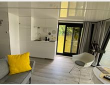 Innenraum eines modernen Hausbootes mit weißer Küchenzeile, grauem Sofa mit gelbem Kissen, weißem Stuhl und großen Glastüren, die einen Blick auf das Wasser und die umliegende Natur ermöglichen. Ein Steuerrad aus Holz befindet sich neben den Türen, und der Raum ist mit hellgrauem Laminatboden ausgestattet.