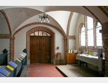 Innenraum einer Kirche mit gotischen Spitzbögen aus Backstein, einem Holztürportal und gewölbter Decke. Links stehen Kirchenbänke mit ausgelegten Broschüren, rechts ein Tisch mit Büchern und Prospekten vor Buntglasfenstern.