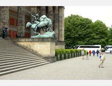 Treppenaufgang eines historischen Gebäudes mit ionischen Säulen, daneben eine Bronzestatue eines Reiters im Kampf mit einem Löwen. Im Hintergrund Busse, Spaziergänger und grüne Bäume.