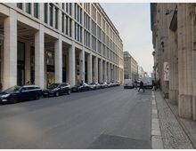 Straßenansicht in einer urbanen Umgebung mit modernen Gebäuden, die von breiten Säulen geprägt sind. Entlang der Straße parken Autos, und auf dem Gehweg sind einige Fußgänger sowie Lieferwagen zu sehen.