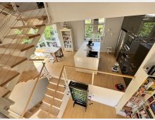 Offener, moderner Wohn- und Küchenbereich über zwei Ebenen mit Holztreppe, Geländern aus Drahtseilen und hellen Holzböden. Große Fenster, eine zentrale Kücheninsel, ein Esstisch sowie Einbauschränke und Regale prägen den lichtdurchfluteten Raum.