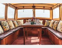 Innenansicht eines rustikalen Bootes mit Lederbänken, die mit bunten Kissen im floralen Design dekoriert sind. In der Mitte steht ein Holztisch, darauf befinden sich Gläser und kleine dekorative Blumenvasen; die Fenster sind mit durchsichtigen Planen bedeckt.