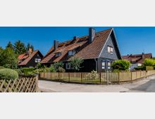 Einfamilienhaus aus dunklem Holz mit rotem Satteldach, umgeben von einem gepflegten Garten und einem Holzzaun; im Hintergrund weitere ähnlich gestaltete Häuser und Bäume unter klarem, blauem Himmel.