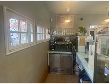 Kleine Café-Theke mit einer grünen Espressomaschine, Gläsern und Tassen auf einem Regal sowie einer Menükarte an der Wand. Im Hintergrund sind hohe Fenster mit weißen Rahmen und draußen blühende Pflanzen sichtbar.