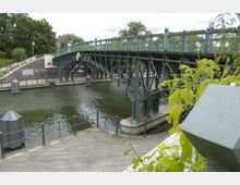 Grüne Metallbrücke mit Gittergeländer über einem Kanal, flankiert von Gehwegen und begrünten Uferbereichen. Im Vordergrund befinden sich Pflastersteine und Geländer; im Hintergrund sind Bäume und ein bewölkter Himmel zu sehen.