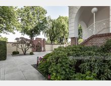 Gepflegter Hof mit einer Gartenanlage, flankiert von einer niedrigen Backsteinmauer und Bäumen. Rechts ist eine geschwungene Treppe mit verziertem Geländer an einem Gebäude aus Sichtziegeln, während vorne ein kleiner gepflegter Strauch und blühende Hortensien sichtbar sind.