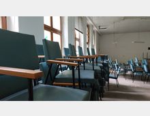 Ein leerer Raum mit gestapelten Stühlen, die gepolsterte Sitzflächen und Armlehnen aus Holz haben. Große Fenster mit Holzrahmen lassen Tageslicht herein, während die Decke mit schlichten Hängelampen ausgestattet ist.