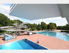 Ein Freibad mit mehreren Schwimmbecken, einer großen Wasserrutsche aus Metall und umliegender gepflasterter Fläche. Im Hintergrund sind Bäume und ein parkähnliches Gelände erkennbar.