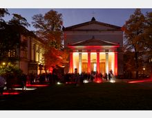 Neoklassizistische Kirche mit Säulenportikus, beleuchtet in roten und gelben Farben bei Dämmerung. Vor der Kirche versammeln sich Menschen, umgeben von herbstlich gefärbten Bäumen und beleuchteten Gehwegen.