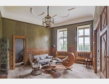 Wohnraum mit historischem Charme, geprägt von Holzwänden, einem grünen Kachelofen, großen Sprossenfenstern und einer hohen Stuckdecke. Das Mobiliar umfasst moderne und klassische Elemente wie einen Ledersessel, ein Sofa, einen Holzschaukelstuhl und einen runden Tisch auf einem Fellteppich.