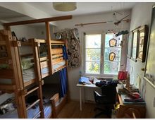 Ein Kinderzimmer mit einem Etagenbett aus Holz links, einem Schreibtisch mit Stuhl rechts vor einem Fenster mit Blick auf Bäume. An der Wand hängen Bilder, Dekorationen sowie ein Mobile, und auf dem Schreibtisch liegen Bücher und Bastelmaterialien.