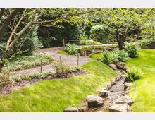 Gepflegter Garten mit einem geschwungenen Weg, gesäumt von niedrigen Seilgeländern und grüner Vegetation. Ein kleiner, von Steinen eingerahmter Bach fließt durch die Rasenflächen, umgeben von Farnen und Bäumen.