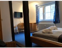 Ein einfach eingerichtetes Schlafzimmer mit einem Doppelbett aus Korbgeflecht, gerollten Handtüchern darauf, einem großen Fenster mit blauen Vorhängen, einer Wandheizung darunter und einem an der Wand montierten Fernseher. Im Hintergrund stehen ein Korbsessel und ein kleiner Beistelltisch.