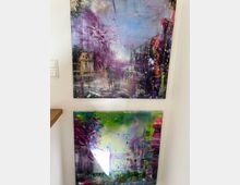 Zwei abstrakte Gemälde an einer weißen Wand, oben ein Bild mit violetten, blauen und goldenen Farbtönen, das eine Stadtlandschaft mit Personen und Gebäuden andeutet, und unten ein Werk in grünen und violetten Tönen, das eine natürliche Landschaft mit Bäumen darstellt. Links ist ein Teil einer Holzablage sichtbar.