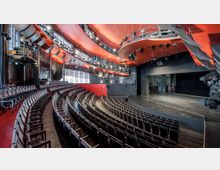 Halbrundes Theater mit ansteigender Sitzplatzanordnung, einer großen Bühne und einem modernen roten Deckenelement mit integrierter Beleuchtung und Technik. Die Glaswand im Hintergrund lässt Tageslicht in den Raum.