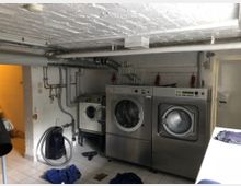 Waschküche mit weißen Wänden und Fliesenboden. Zu sehen sind mehrere Waschmaschinen und Trockner aus Edelstahl sowie Rohre an der Decke; ein Staubsauger und Wäsche liegen im Vordergrund.