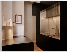 Moderne Küche mit matt schwarzen Schrankfronten und einer durchgehenden Edelstahl-Arbeitsfläche samt Spüle, Rückwand und Oberschränken. Links steht ein Edelstahlblock mit Wandlampe und gerahmtem Bild vor hellen, glatten Wänden; der Boden besteht aus Holzdielen.