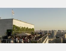Dachterrasse eines Gebäudes mit grüner Wandbegrünung, Sonnenschirmen und einer großen Anzahl von Menschen, die sich unterhalten. Im Hintergrund ist eine städtische Skyline mit modernen und historischen Gebäuden zu sehen, bei klarem Himmel und tiefstehender Sonne.