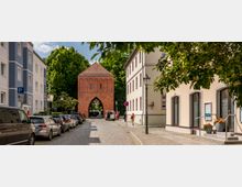 Eine gepflasterte Straße mit parkenden Autos führt zu einem großen mittelalterlichen Torbogen aus rotem Backstein, umgeben von Bäumen. Auf beiden Seiten der Straße stehen moderne Gebäude mit Putzfassaden.