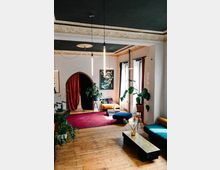 Wohnzimmer in einem Altbau mit einem Holzboden, verziertem Deckenstuck und dunkelgrüner Decke. Der Raum ist eingerichtet mit Pflanzen, bunten Möbeln, einem roten Teppich und einer modernen, hängenden Beleuchtung.