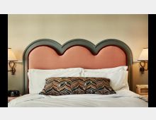 Ein Bett mit einem auffälligen Kopfteil in Herzform, bezogen mit rosa Stoff und einem dunkelgrünen Rahmen. Auf dem Bett liegen weiße Kissen und eine dekorative Kissenrolle mit gemustertem Stoff, eingerahmt von zwei Wandlampen auf beige Wänden.