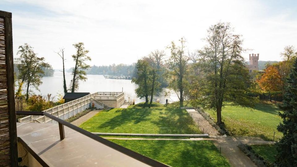 Blick von einem Balkon über eine gepflegte Grünanlage mit Wegen, Bäumen und einer Terrassenplattform hin zu einem See. Im Hintergrund sind das Ufer mit Bootsanlegern und ein turmartiges Gebäude zwischen den Bäumen zu sehen.