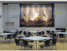 Ein Veranstaltungsraum mit mehreren leeren Tischen und schwarzen Stühlen auf einem Holzfußboden. An der Stirnwand befindet sich ein großes Wandbild einer Flugzeugnase mit Propellern, darüber steht „Flugplatz Schönghage“.